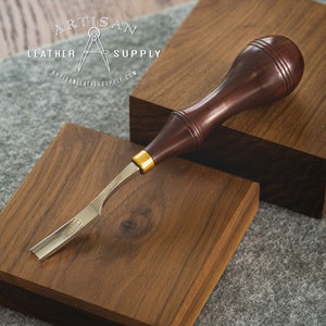 May include: A close-up of a wood carving tool with a brown wooden handle and a metal blade. The tool is resting on a wooden surface. The text "ARTISAN LEATHER SUPPLY" is visible in the background.