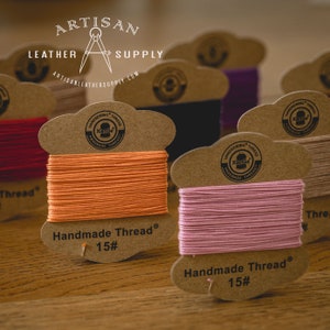 May include: Close-up of two spools of orange and pink waxed thread. The spools are labeled "Handmade Thread® 15#" and feature a brown paper label with a logo that says "HANDSEWING THREAD XianGe HANDMADE THREAD".