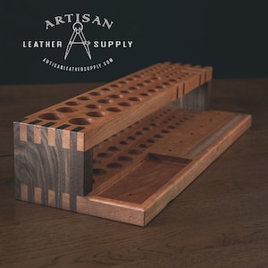 Puede incluir: Un organizador de madera de dos niveles con una base de nogal y una parte superior de cerezo. El nivel superior tiene 36 agujeros para guardar objetos pequeños, mientras que el nivel inferior tiene un compartimento más grande y una bandeja más pequeña. El organizador está etiquetado como "Artisan Leather Supply" con un logotipo de brújula.