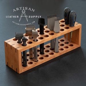 May include: A rectangular light brown wooden tool organizer with multiple circular holes for storage. Several metal leatherworking tools with various head shapes and dark handles are inserted into the slots. The text ARTISAN LEATHER SUPPLY is visible.
