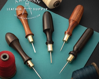 May include: Five leather stitching awls with wooden handles and gold metal tips. The awls are arranged on a teal blue surface with spools of thread in the background. The text "Artisan Leather Supply" is visible in the top left corner of the image.