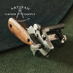 May include: A silver leather working tool with a wooden handle. The tool has a ruler, a rotating blade, and a knob for adjusting the blade depth. The tool is on a green fabric surface. The text "ARTISAN LEATHER SUPPLY" is visible in the top left corner of the image.