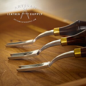 May include: Three leather working tools with wooden handles and metal blades. The tools are arranged in a row on a wooden surface. The tools are labeled "Artisan Leather Supply".
