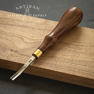 May include: A close-up of a wood carving tool with a brown wooden handle and a brass ferrule. The blade is curved and has a Japanese inscription on it. The tool is resting on a wooden surface.