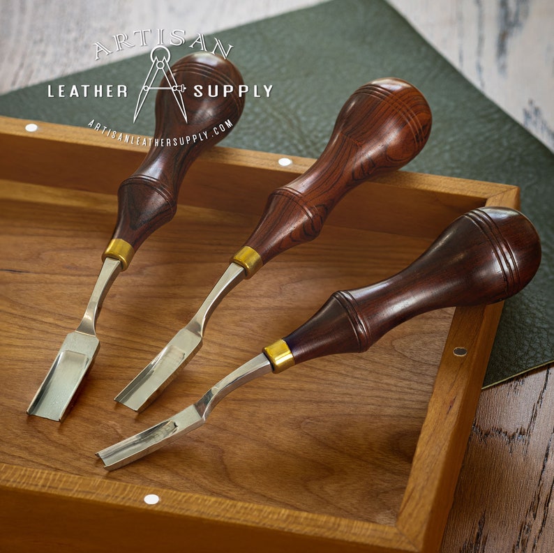 May include: Three leather working tools with wooden handles and metal blades. The tools are in a wooden box with the text "Artisan Leather Supply" on the top.