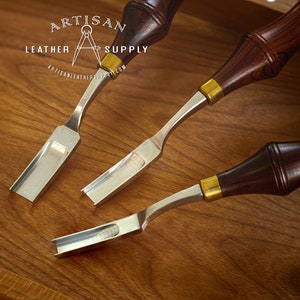 May include: Three leather working tools with wooden handles and metal blades. The tools are arranged on a wooden surface. The tools are labeled "Artisan Leather Supply".