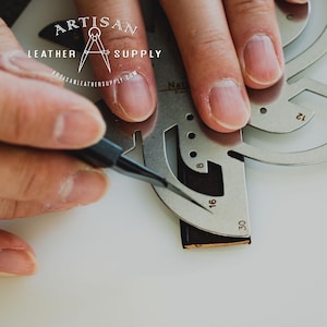May include: A metal leather crafting tool with markings for different sizes. The tool is being used to trace a line on a piece of brown leather.