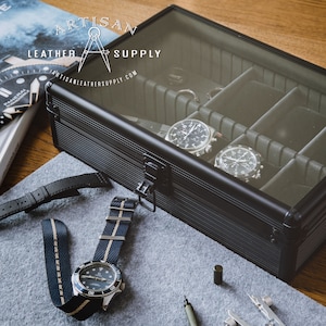 May include: A black watch case with a clear lid, holding two watches. A watch with a blue and beige strap and a black dial sits on a gray surface. The Artisan Leather Supply logo is visible.