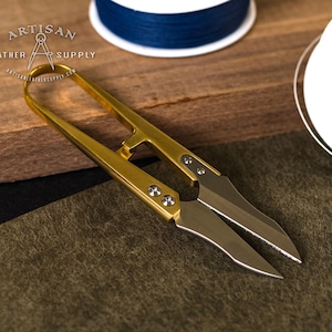 May include: Gold-toned leather shears with a pointed tip and a small loop at the top. The shears are on a brown and green surface.
