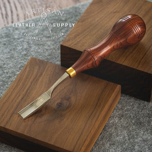 May include: A close-up of a leather working tool with a wooden handle and a metal blade. The tool is resting on a wooden surface. The handle is a dark reddish brown with a wood grain pattern. The blade is silver and has a slight curve. The tool is labeled "Artisan Leather Supply".