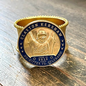 May include: A gold-coloured metal napkin ring with a round, raised, gold-coloured metal medallion featuring a baseball player in a blue and white uniform with the word "Dodgers" on the front. The medallion has the text "Clayton Kershaw", "June 18, 2014", "No-Hitter vs. Colorado" and stars around the edge.