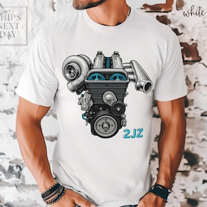May include: White t-shirt featuring a detailed illustration of a 2JZ engine in black, gray, and blue. The text "2JZ" is printed in blue. The shirt has the text "SHIPS NEXT DAY" in the upper left corner and the word "white" in the upper right corner.