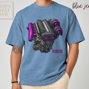 May include: Blue t-shirt featuring a detailed illustration of an engine with purple accents. The text "RB26" is printed in pink. The words "blue jean" are written in cursive. The shirt has a relaxed fit.