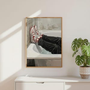 May include: A framed artwork depicts a person's legs in a bathtub. The person wears black pants and red and white sneakers. The artwork is in a gold frame and is displayed on a white surface.