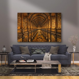 May include: A canvas print showing a library interior with arched ceilings and rows of bookshelves. The artwork uses warm, golden tones and a reflective surface, creating depth. The print hangs above a blue sofa. The room has a rug and two side tables.