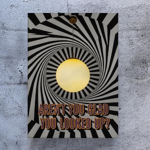 Puede incluir: Un póster con un diseño en espiral en blanco y negro, una lámpara esférica amarilla y el texto "AREN'T YOU GLAD YOU LOOKED UP?" en morado y amarillo. La lámpara cuelga.