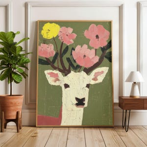 May include: A framed art print featuring a deer with antlers replaced by pink and yellow flowers. The deer is painted in cream and brown tones against a green background. The artwork is displayed in a room with a plant and a lamp.