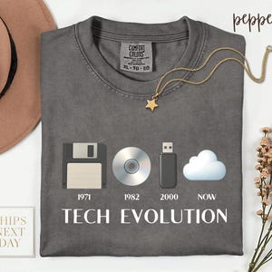 May include: A gray t-shirt with the words "TECH EVOLUTION" in white. The shirt features a graphic of a floppy disk, CD, USB drive, and cloud, with the years 1971, 1982, 2000, and NOW below each image.
