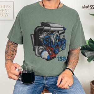 May include: A moss green t-shirt featuring a detailed illustration of an engine with blue, black, and red components. The text "13B" is printed in blue on the lower right. The shirt has a relaxed fit.