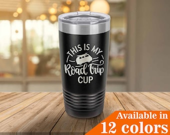 May include: Black travel mug with a silver rim and lid. The mug features the text "THIS IS MY Road Trip CUP" in white, along with a graphic of a camper van. The mug is on a wooden surface. Available in 12 colors.