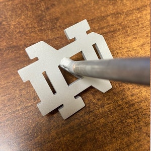 May include: A silver metal cutout of the letters "ND" with a silver metal rod going through the center of the letters.