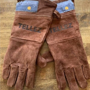 Op de afbeelding: Een paar bruine leren ovenwantjes met de tekst "Outset" en "Tellez" erop gedrukt. De wantjes zijn ontworpen voor gebruik met een grill.