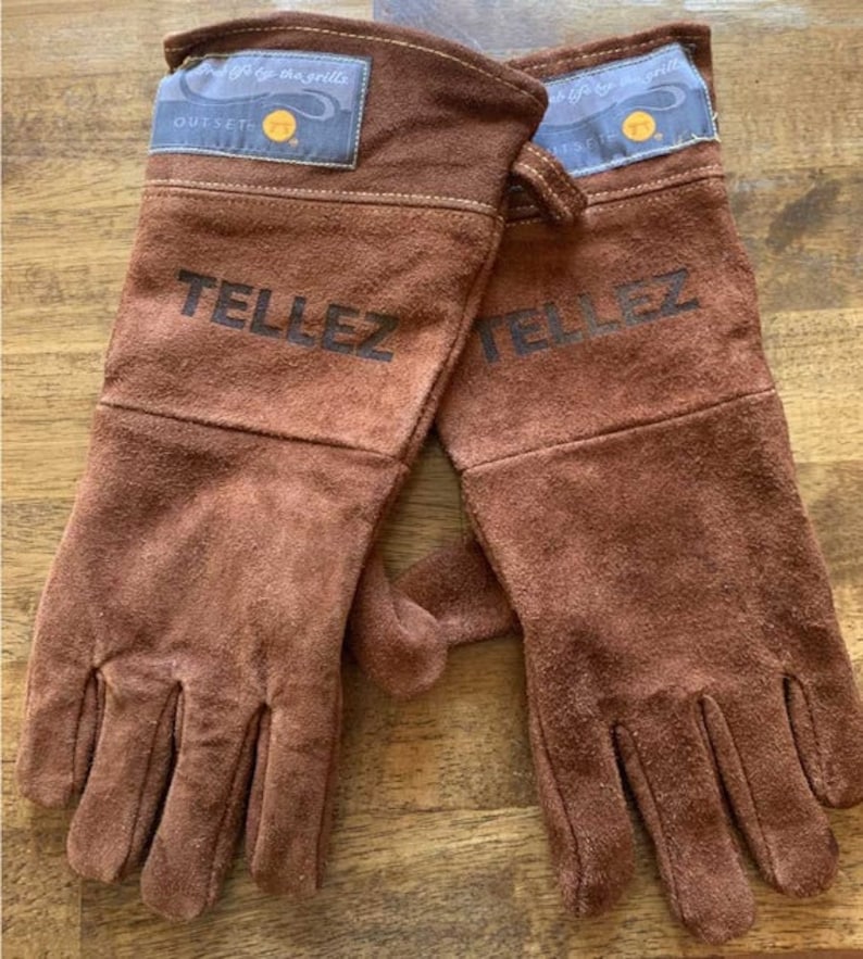 Op de afbeelding: Een paar bruine leren ovenwantjes met de tekst "Outset" en "Tellez" erop gedrukt. De wantjes zijn ontworpen voor gebruik met een grill.