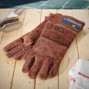 Op de afbeelding: Een paar bruine leren grillhandschoenen met de tekst "DCL" geborduurd op de achterkant van de linker handschoen. De handschoenen hebben een bruine leren band met de tekst "Just try to outgrill" en een logo op de achterkant van de rechter handschoen.