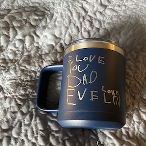 May include: A navy blue insulated mug with a silver rim and handle. The mug has the handwritten text "I LOVE YOU DAD EVELYN" in a silver color. The mug is sitting on a gray quilted surface.