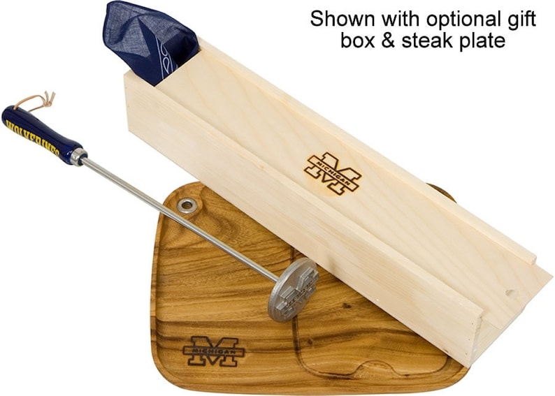 May include: A wooden box with a blue bandana inside, a wooden steak board with a metal steak pounder, and a metal skewer with a blue and yellow handle. The box and board are both engraved with the University of Michigan logo.