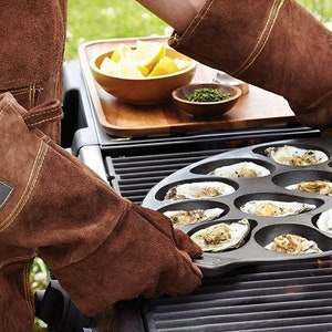 Op de afbeelding: Een persoon met bruine leren grillhandschoenen houdt een gietijzeren oestergrillpan vast met 12 individuele compartimenten. De pan staat op een gasgrill met zichtbare vlammen eronder.