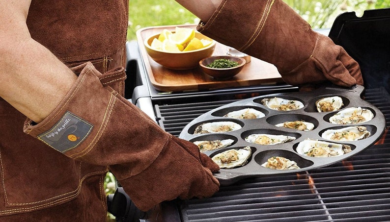 Op de afbeelding: Een persoon met bruine leren grillhandschoenen houdt een gietijzeren oestergrillpan vast met 12 individuele compartimenten. De pan staat op een gasgrill met zichtbare vlammen eronder.