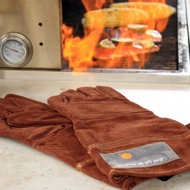 Op de afbeelding: Twee bruine leren grillhandschoenen met een gele gestikte naad en een zwart label met de tekst "Black Rifle the Smoke Outfitters".