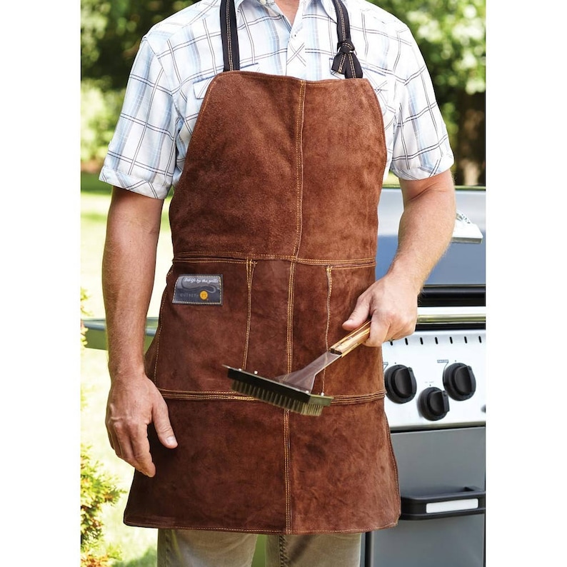 Op de afbeelding: Een bruine leren schort met meerdere zakken en een gereedschapshouder. Het schort heeft gele stiksels en een klein patch met de tekst "Design for the grill".