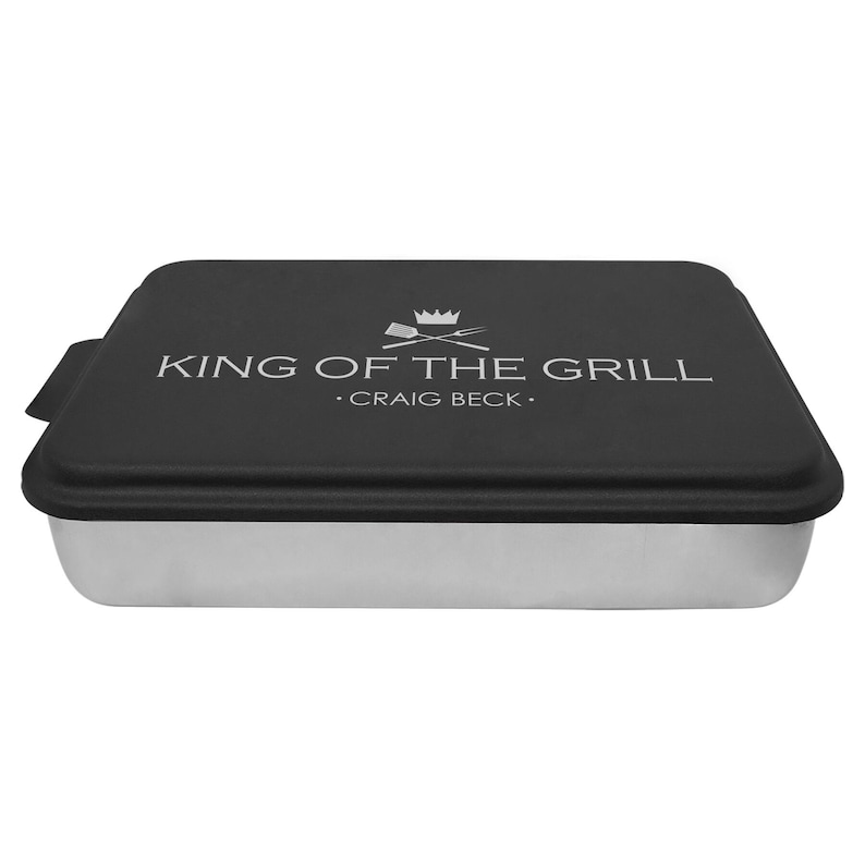 May include: A black and silver baking pan with a lid. The lid has a white graphic that says "King of the Grill" with a crown and crossed grilling tools. The text below the graphic says ".Craig Beck."