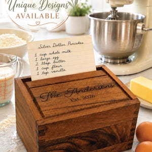 May include: A rectangular dark brown wooden recipe box with finger-jointed corners. The lid is engraved with the text "The Andersons Est. 2026". A white recipe card titled "Silver Dollar Pancakes" with handwritten ingredients is slotted into the lid.