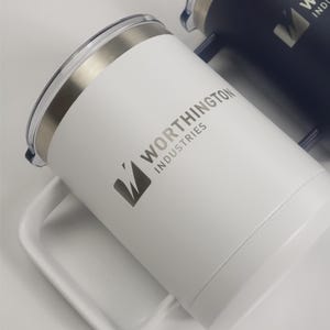May include: White insulated travel mug with a silver rim and handle. The mug features the text "WORTHINGTON INDUSTRIES" and a logo in gray. A second, dark blue mug is partially visible in the background.