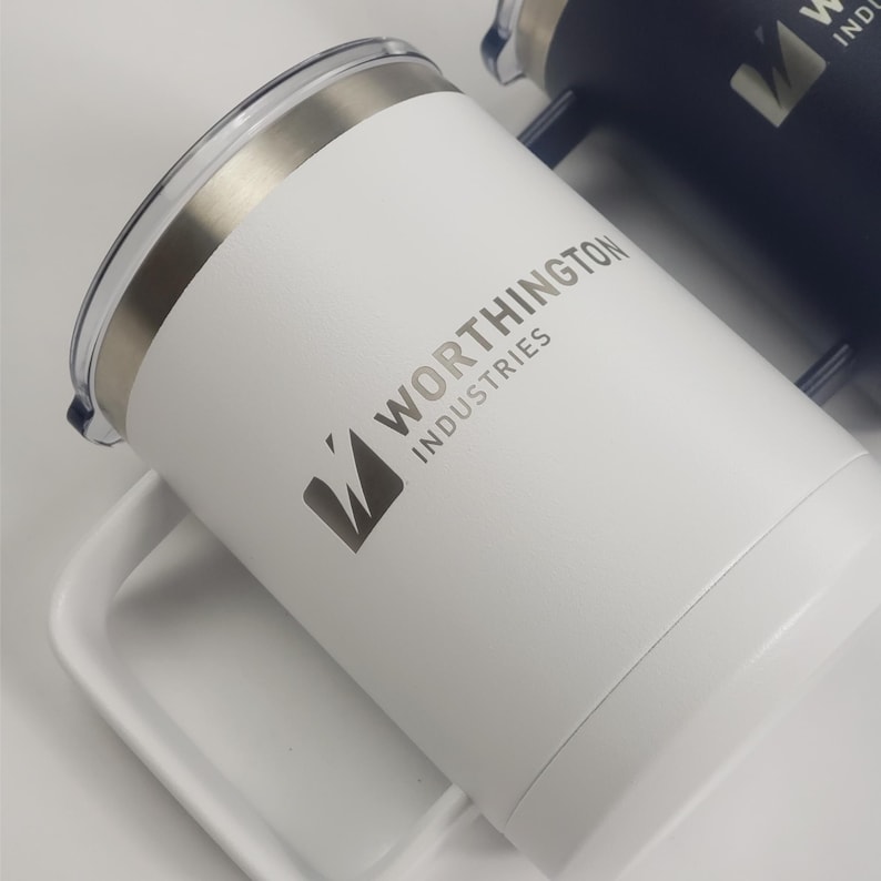 May include: White insulated travel mug with a silver rim and handle. The mug features the text "WORTHINGTON INDUSTRIES" and a logo in gray. A second, dark blue mug is partially visible in the background.