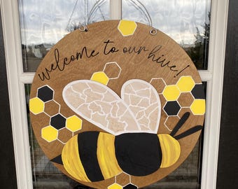 May include: Round wooden welcome sign with a bee design. The sign features a large bee with yellow and black stripes, honeycomb patterns, and the text "welcome to our hive!" in cursive.