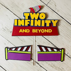 May include: A red, yellow, and blue cake topper with the text "TWO INFINITY AND BEYOND" and a small rocket ship. Two purple and green striped cake toppers with a yellow border.