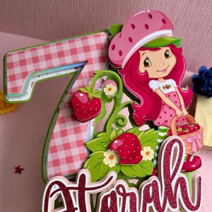 May include: A pink and green cake topper featuring a cartoon strawberry character with red hair, a pink hat, and a white dress. The character is holding a basket of strawberries. The cake topper also includes the number 7 and the name "Starla" in pink glitter letters.