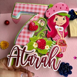 May include: A pink and green cake topper featuring a strawberry shortcake character, the number 7, and the name "Airah" in red glitter. The topper is decorated with strawberries, leaves, and flowers.