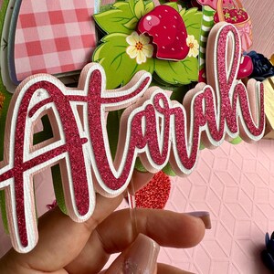 May include: Pink glitter cake topper with the name "Airah" written in a cursive font. The topper is on a clear plastic stick.