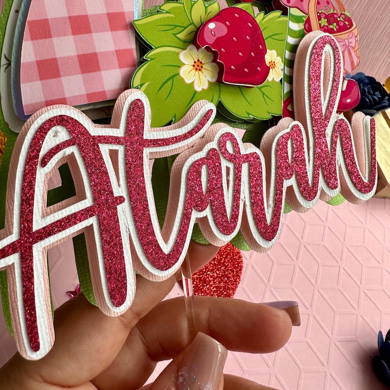 May include: Pink glitter cake topper with the name "Airah" written in a cursive font. The topper is on a clear plastic stick.