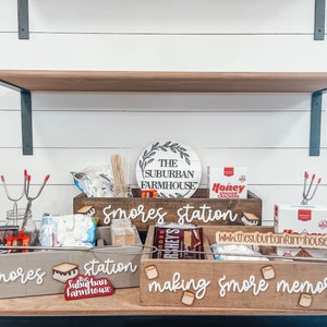 May include: A wooden box with the words "Smores station" written on it. The box is filled with various ingredients for making s'mores, including graham crackers, marshmallows, and chocolate. There is also a sign that says "The Suburban Farmhouse" and a sign that says "Making smore memories".