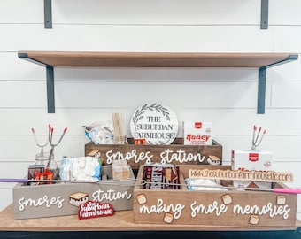 May include: A wooden box with the words "Smores station" written on it. The box is filled with various ingredients for making s'mores, including graham crackers, marshmallows, and chocolate. There is also a sign that says "The Suburban Farmhouse" and a sign that says "Making smore memories".
