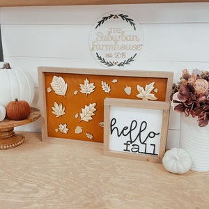 May include: A wooden sign with the text "The Suburban Farmhouse est. 2018" surrounded by a wreath. A white wooden sign with the text "hello fall" is next to a framed piece of art with a brown background and gold leaf cutouts. There are white and brown pumpkins on a wooden pedestal.