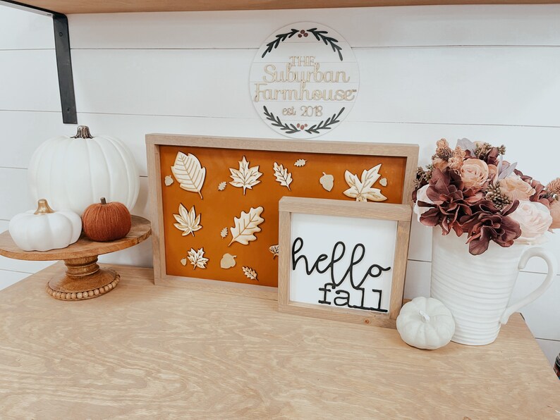May include: A wooden sign with the text "The Suburban Farmhouse est. 2018" surrounded by a wreath. A white wooden sign with the text "hello fall" is next to a framed piece of art with a brown background and gold leaf cutouts. There are white and brown pumpkins on a wooden pedestal.