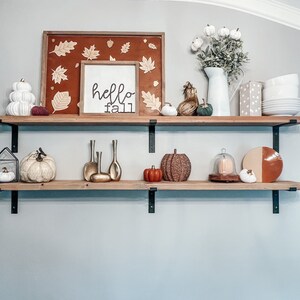 May include: Two wooden shelves with black metal brackets, decorated with fall-themed items, including pumpkins, gourds, a sign that says "hello fall", and a framed picture with fall leaves.