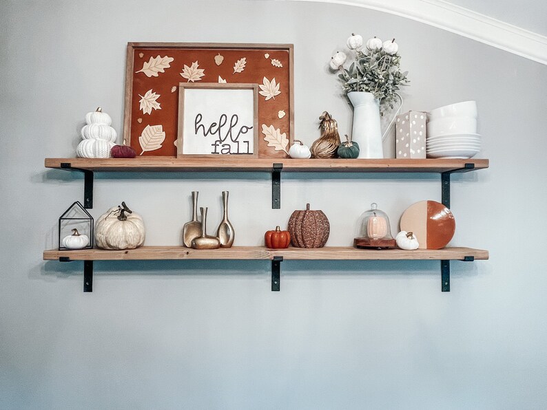May include: Two wooden shelves with black metal brackets, decorated with fall-themed items, including pumpkins, gourds, a sign that says "hello fall", and a framed picture with fall leaves.